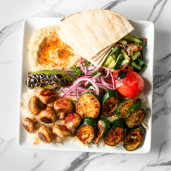 Mediterranean platter with hummus, pita, cucumber salad, grilled vegetables, and mushrooms on a white plate.