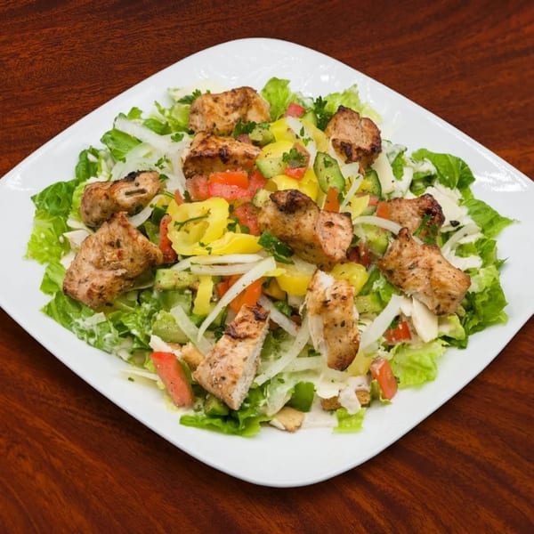 A Mediterranean chicken salad with lettuce, tomatoes, cucumbers, onions, and yellow peppers on a white plate.